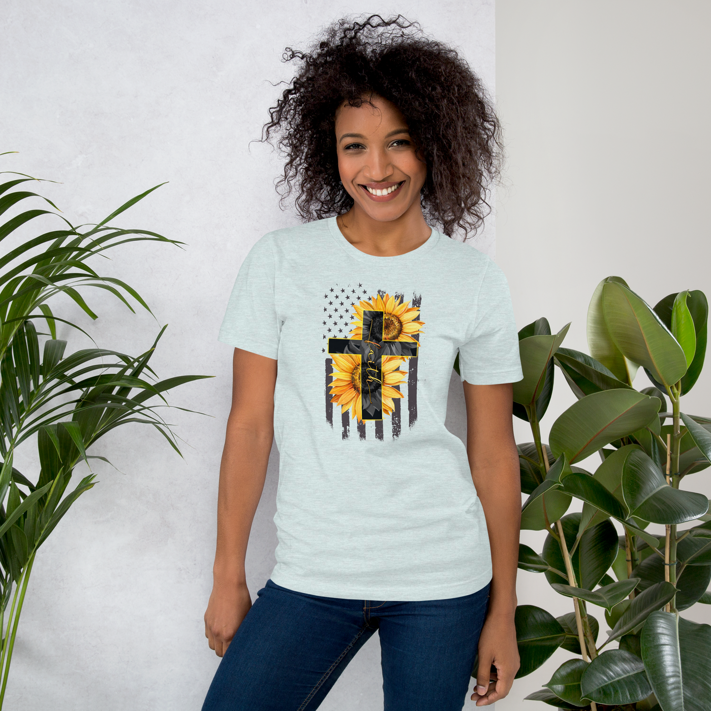 Woman wearing a light blue t-shirt with a sunflower and cross design, standing between two plants.